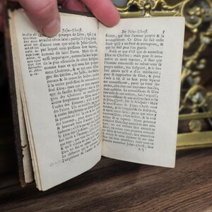 K&ouml;nnte beinhalten: Ein altes Buch mit vergilbten Seiten ist ge&ouml;ffnet und zeigt Text in einer Fremdsprache. Das Buch hat einen dunkelbraunen Einband, und eine Hand h&auml;lt die Seiten offen. Im Hintergrund ist ein dekoratives Messingobjekt zu sehen. Der Text enth&auml;lt "Jesus-Chrift".