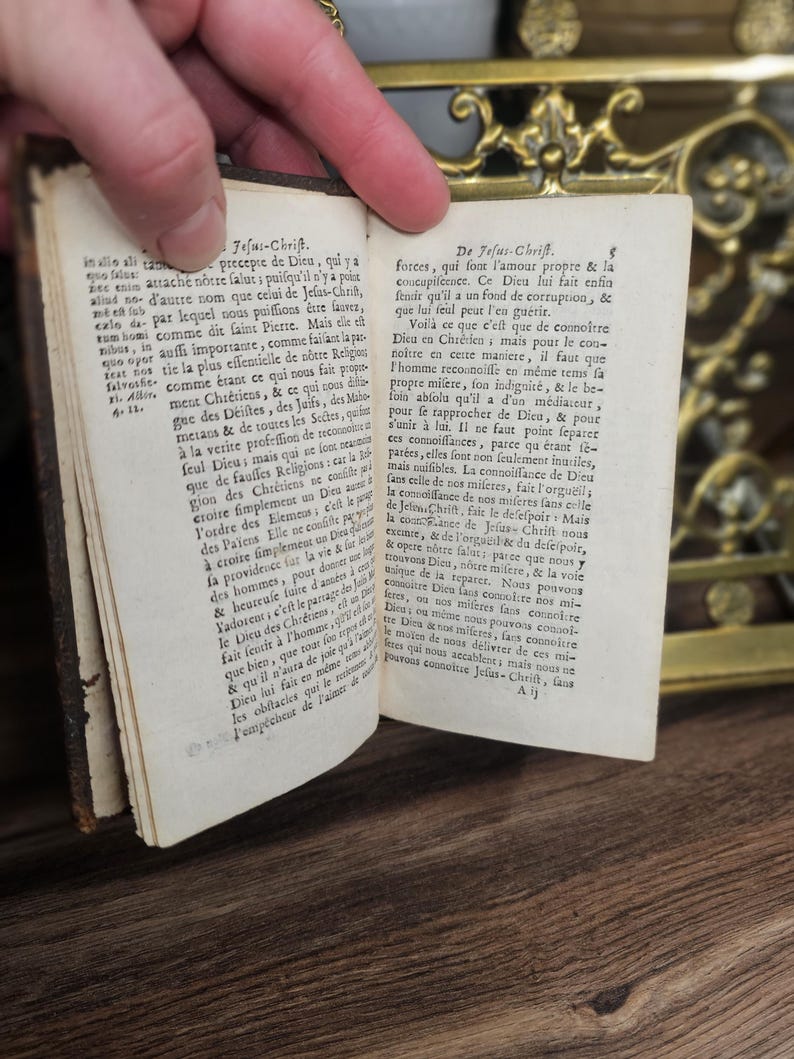 K&ouml;nnte beinhalten: Ein altes Buch mit vergilbten Seiten ist ge&ouml;ffnet und zeigt Text in einer Fremdsprache. Das Buch hat einen dunkelbraunen Einband, und eine Hand h&auml;lt die Seiten offen. Im Hintergrund ist ein dekoratives Messingobjekt zu sehen. Der Text enth&auml;lt "Jesus-Chrift".