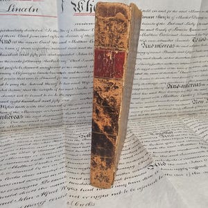 May include: An old book with a distressed leather cover. The spine has a red panel with an embossed design. The book is placed on a background of aged documents with handwritten text, suggesting historical significance.