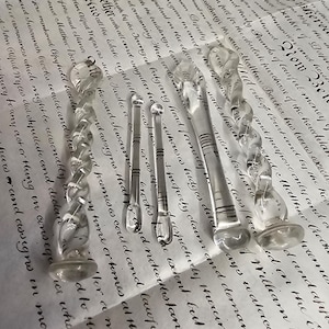May include: Several clear glass dip pens with decorative spiral and bulbous designs are displayed. The pens of varying sizes are arranged on a vintage document with handwritten script.