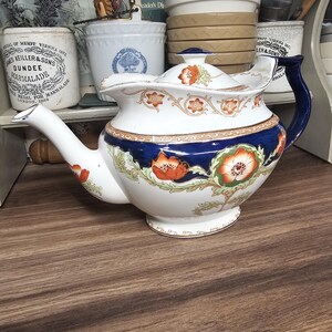 Può includere: Una teiera in ceramica bianca e blu con un motivo floreale. La teiera ha un beccuccio curvo, un manico blu e un coperchio con un piccolo pomello. È decorata con fiori arancioni e verdi e si trova su una superficie di legno.