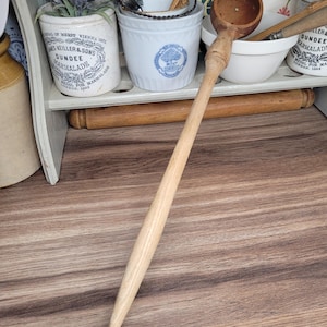 May include: A long-handled wooden ladle with a rounded bowl. The ladle is light-colored and appears to be made of a single piece of wood. The background includes white ceramic containers and a shelf with vintage-style items.