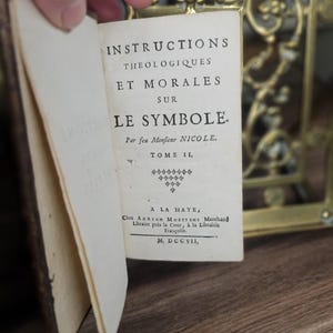 K&ouml;nnte beinhalten: Ein altes Buch mit dem Titel "INSTRUCTIONS THEOLOGIQUES ET MORALES SUR LE SYMBOLE" ist ge&ouml;ffnet und zeigt franz&ouml;sischen Text. Die Seiten des Buches sind gealtert, und der Einband ist dunkelbraun. Der Text ist in schwarzer Schrift.