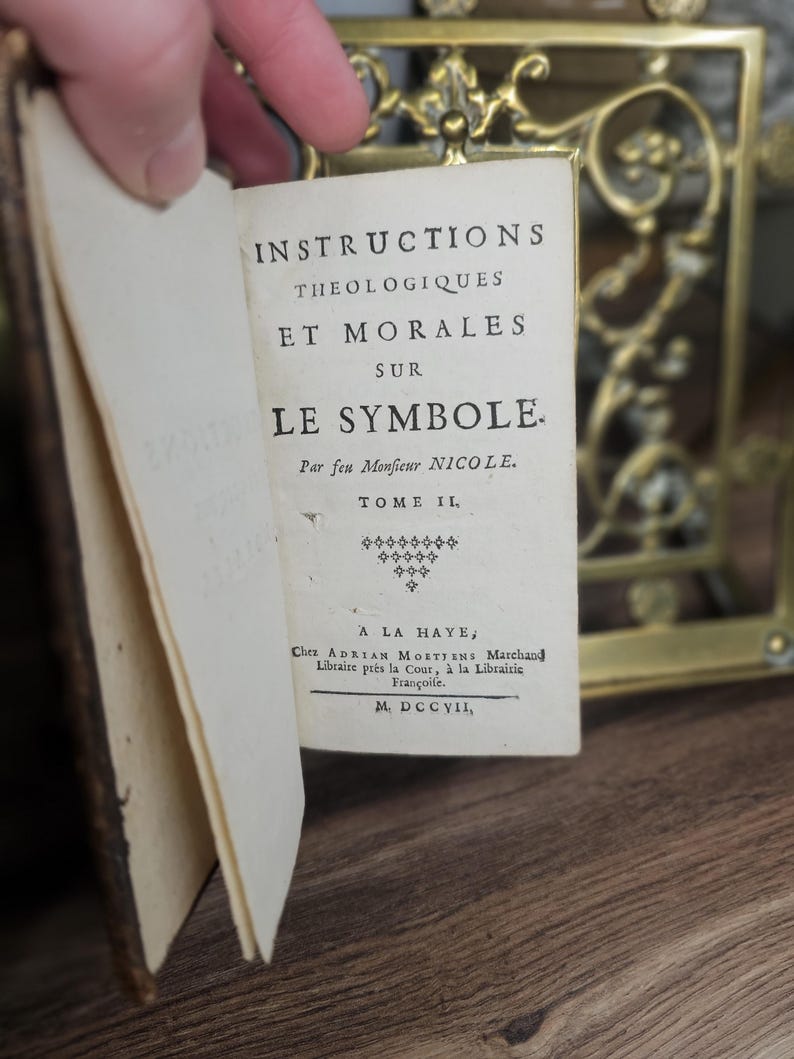 K&ouml;nnte beinhalten: Ein altes Buch mit dem Titel "INSTRUCTIONS THEOLOGIQUES ET MORALES SUR LE SYMBOLE" ist ge&ouml;ffnet und zeigt franz&ouml;sischen Text. Die Seiten des Buches sind gealtert, und der Einband ist dunkelbraun. Der Text ist in schwarzer Schrift.