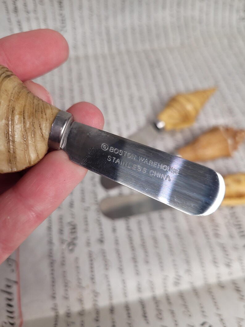 May include: A set of three small, wooden, seashell-shaped butter knives. The knives have stainless steel blades with the text "BOSTON WAREHOUSE STAINLESS CHINA" printed on the blade.