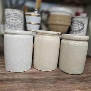 Può includere: Tre vasi cilindrici in ceramica disposti in fila. Il vaso a sinistra è bianco, quello centrale è color crema chiaro e quello a destra è di un beige tenue. Ogni recipiente presenta un bordo spesso e arrotondato e una finitura smaltata.