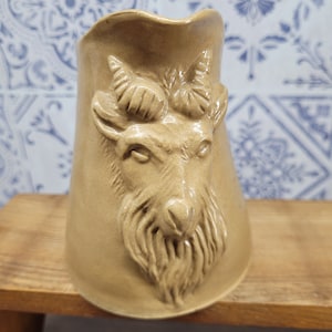 May include: A ceramic pitcher shaped like a goat's head. The pitcher is a light brown color and has a detailed design of the goat's face, including its horns, eyes, nose, and beard.