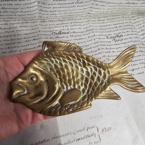 May include: A vintage brass fish-shaped decorative object. The fish has detailed scales, fins, and an open mouth. The brass has a warm, aged patina. The object is likely a decorative piece or a small tray.
