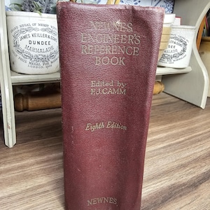 May include: A maroon-colored book titled "Newnes Engineer's Reference Book" with gold lettering. The book is edited by F.J. Camm and is the eighth edition. The publisher's name, Newnes, is printed at the bottom. The book is standing upright.