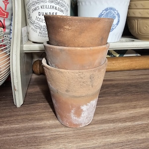 Peut inclure: Trois pots de fleurs en terre cuite empilés sur une surface en bois. Les pots sont de tailles variées et présentent des signes d'usure. Les pots sont de couleur brun chaud et terreux. En arrière-plan, plusieurs récipients en céramique anciens.