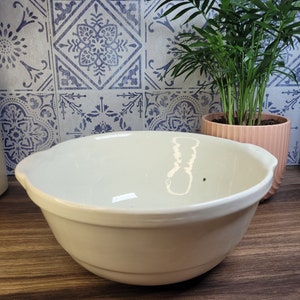 May include: A large, white ceramic mixing bowl with a slightly rounded bottom and a smooth, glossy finish. The bowl is sitting on a wooden surface.