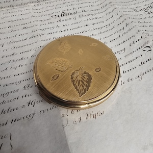 Peut inclure: Un compact rond doré avec un motif de feuilles sur le couvercle. Le compact est fermé et posé sur un fond de papier vieilli avec une écriture manuscrite. Il s'agit d'un article vintage.