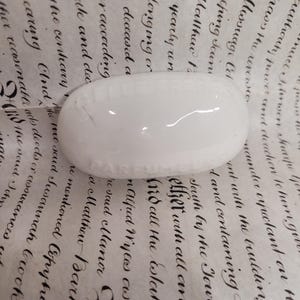 Puede incluir: Un objeto blanco de forma ovalada con una superficie ligeramente texturizada. El objeto parece estar hecho de cerámica o porcelana. El fondo es un papel vintage con escritura manuscrita.