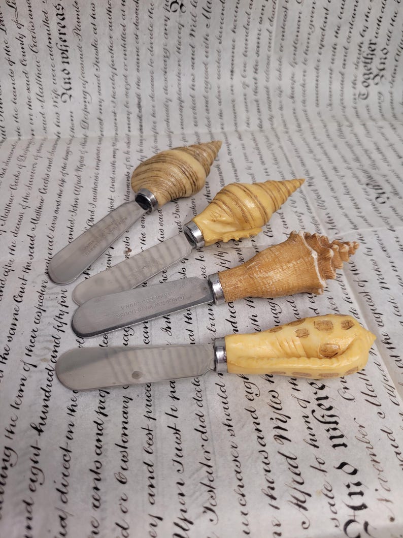 May include: Four silver butter knives with decorative seashell handles. The handles are made of natural seashells with brown and white stripes.