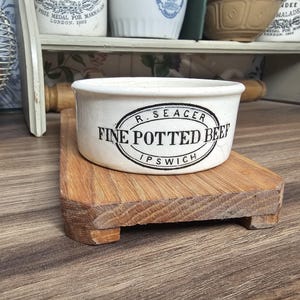 May include: A white ceramic bowl with the text "FINE POTTED BEEF IPSWICH" in black lettering, resting on a small wooden cutting board. The bowl has a black oval border with the name "R. SEACER" above the text. The background includes a shelf with other ceramic items.