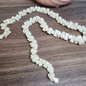 May include: A white carved flower necklace made of bone or ivory. The necklace is made of individual carved flowers that are strung together. The necklace is on a wooden surface.
