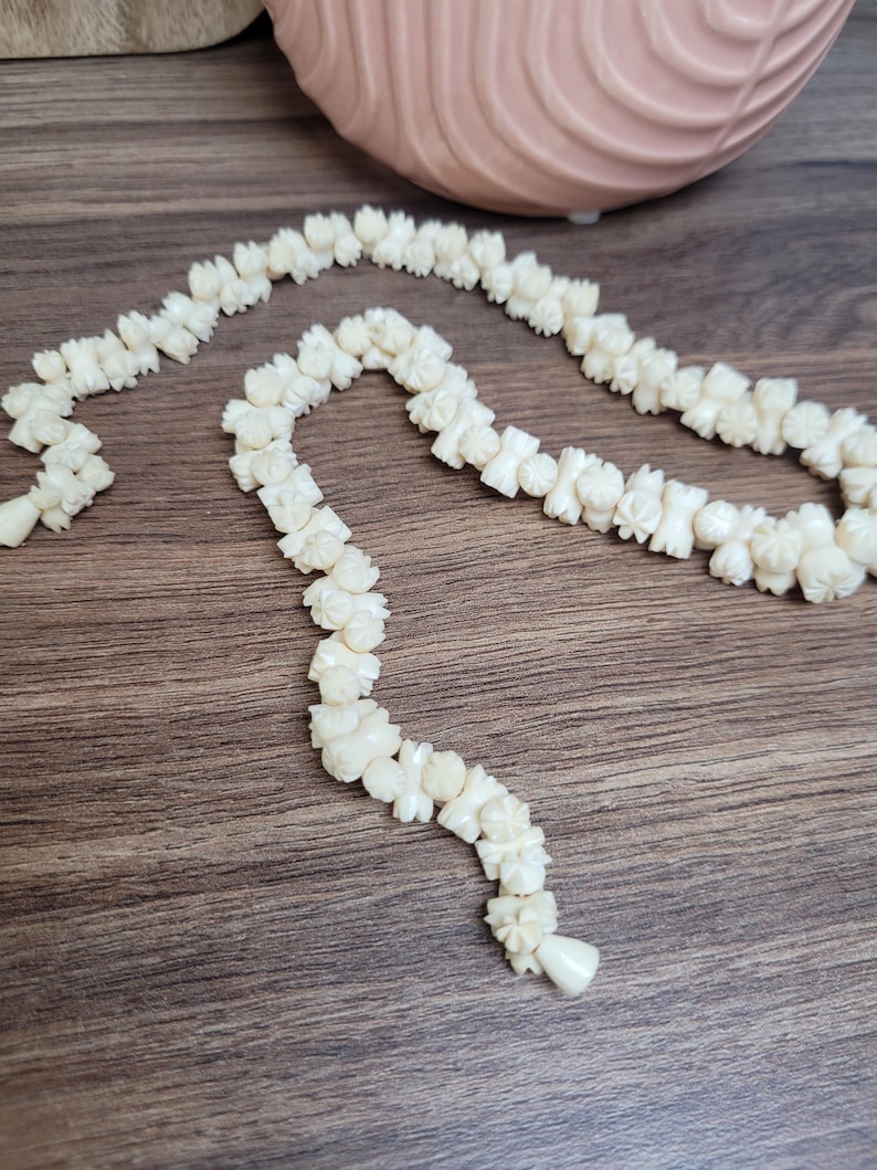 May include: A white carved flower necklace made of bone or ivory. The necklace is made of individual carved flowers that are strung together. The necklace is on a wooden surface.
