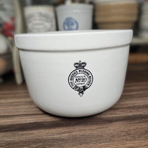 以下が含まれることがあります： 「The Queen's Pudding Boiler No. 20 Challis Patent」と書かれた白黒のエンブレムが付いた白いセラミックプリンボイラー。ボウルは木製の表面にあり、背景には他のセラミックアイテムがあります。