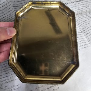 May include: A vintage, octagonal, gold-toned metal tray. The tray has a slightly worn appearance, with visible scratches and a tarnished finish. The edges are raised and angled, and the interior surface is smooth. The tray is held by a hand, with a document in the background.