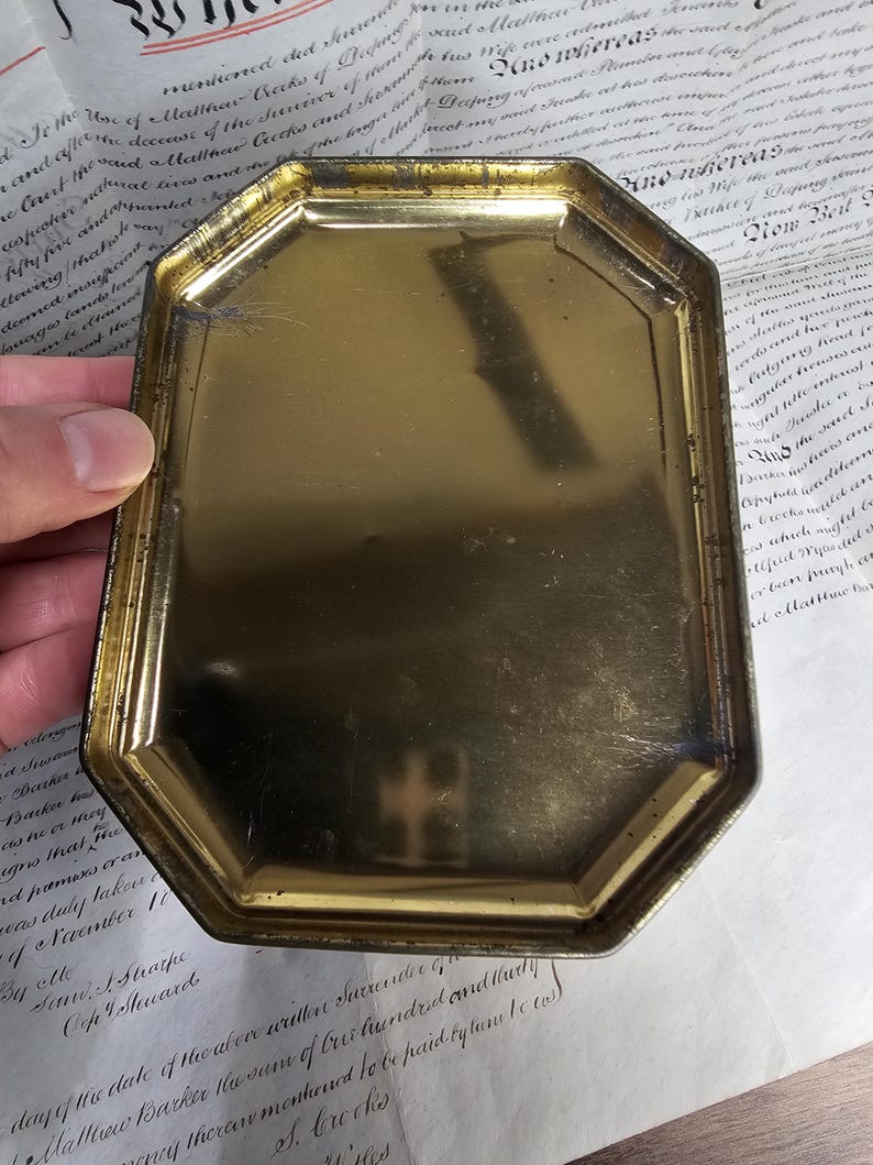 May include: A vintage, octagonal, gold-toned metal tray. The tray has a slightly worn appearance, with visible scratches and a tarnished finish. The edges are raised and angled, and the interior surface is smooth. The tray is held by a hand, with a document in the background.