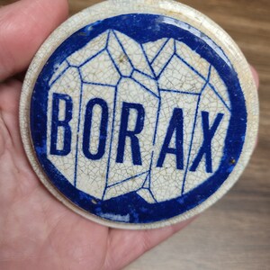 May include: A white ceramic lid with a blue border and a stylized image of crystals. The word "BORAX" is printed in blue letters on the lid.