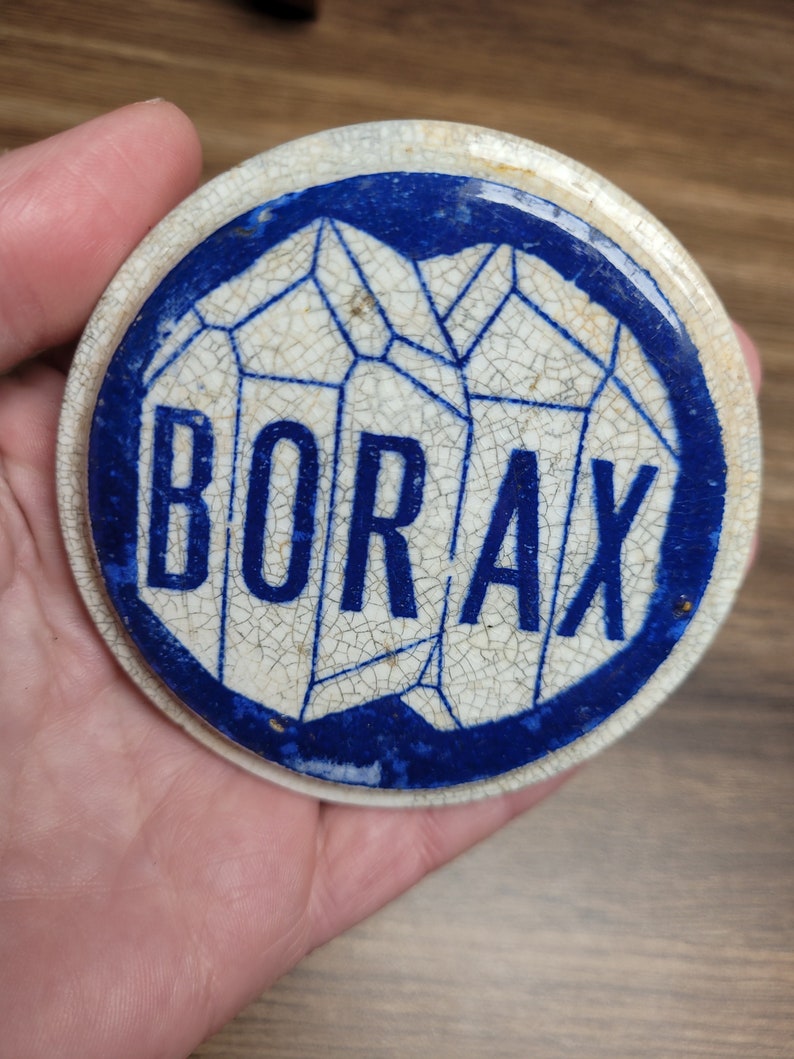 May include: A white ceramic lid with a blue border and a stylized image of crystals. The word "BORAX" is printed in blue letters on the lid.