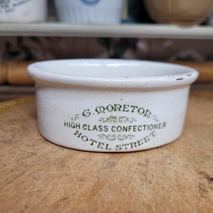 Peut inclure: Un bol en céramique blanc de forme arrondie avec un bord légèrement usé. Le bol porte l'inscription verte "G. MORETON HIGH CLASS CONFECTIONER HOTEL STREET". Le bol est posé sur une surface en bois.