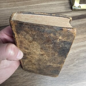 K&ouml;nnte beinhalten: Ein antikes Buch mit einem abgenutzten, braunen Ledereinband. Die Kanten der Seiten sind sichtbar, und das Buch scheint stark benutzt zu sein. Das Leder hat ein geflecktes Aussehen, was auf Alter und Handhabung hindeutet.