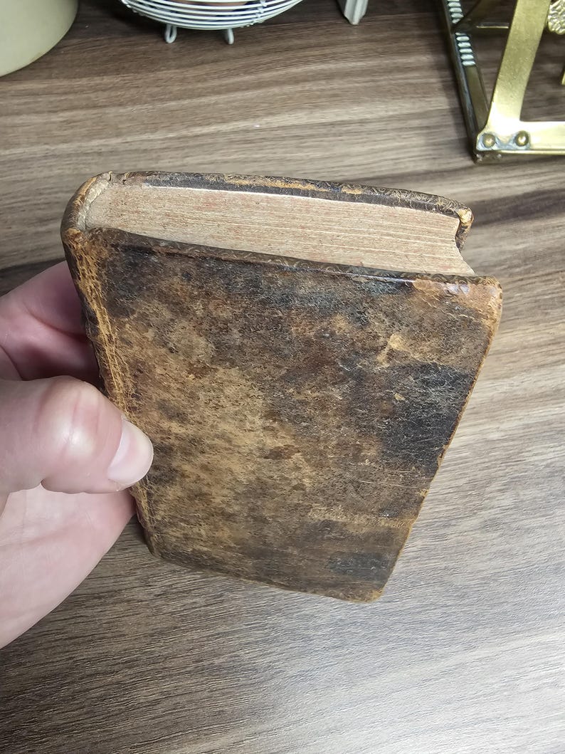 K&ouml;nnte beinhalten: Ein antikes Buch mit einem abgenutzten, braunen Ledereinband. Die Kanten der Seiten sind sichtbar, und das Buch scheint stark benutzt zu sein. Das Leder hat ein geflecktes Aussehen, was auf Alter und Handhabung hindeutet.