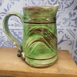 May include: A green ceramic pitcher with brown stripes and a handle. The pitcher has a glazed finish and is sitting on a wooden surface.