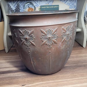 May include: A large, copper planter with an ornate design. The planter has a rounded bottom and a wide, flat rim. The design features four repeating floral motifs.