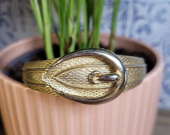 May include: A gold-toned metal bracelet with a buckle fastening. The bracelet is textured and has a slightly worn appearance.