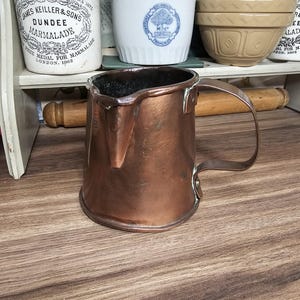 Puede incluir: Una jarra de cobre vintage con asa y boquilla. La jarra es de color marrón rojizo cálido y se encuentra sobre una superficie de madera. Otros elementos en el fondo incluyen un rodillo y tarros. Un artículo de cocina clásico.