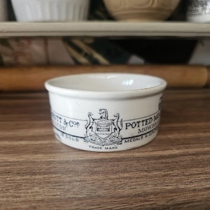 May include: A small, white ceramic bowl with a black and white label wrapped around the exterior. The label reads "POTTED MEAT MOWBRAY & CO LONDON" with a crest and the words "TRADE MARK". The bowl is on a wooden surface.