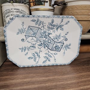 May include: A rectangular ceramic tray with clipped corners, featuring a white base and a blue transferware pattern. The design includes central floral motifs, rose hips, and two geometric box-like structures, all framed by a decorative triangular border.