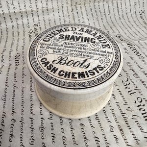 Puede incluir: Un tarro de crema de afeitar vintage con un diseño blanco y negro. El tarro es cilíndrico con tapa, con el texto "Crème D'Amande For Shaving" y "Boots Cash Chemists". El tarro se encuentra sobre un fondo de papel envejecido con escritura manuscrita.