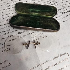 Puede incluir: Gafas antiguas de tipo pince-nez con puente dorado y lentes transparentes, cerca de un estuche ovalado abierto forrado con terciopelo verde. El estuche tiene un exterior plateado. El fondo presenta escritura manuscrita.