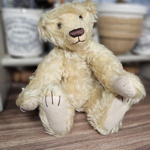 May include: A vintage-style teddy bear with cream-colored fur, a brown embroidered nose, and small black eyes. The bear is sitting with its arms and legs outstretched, showcasing its tan paw pads. A classic plush toy.