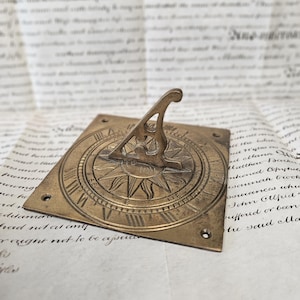 Puede incluir: Un reloj de sol de latón antiguo con una base cuadrada y un gnomon elevado. El reloj de sol presenta números romanos y un diseño de sol. El fondo es un trozo de papel envejecido con texto manuscrito.