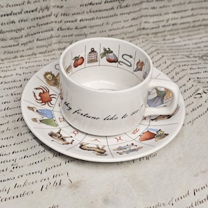 May include: A white ceramic teacup and saucer set featuring colorful illustrations of zodiac signs and symbols. The cup displays the text 'thy fortune like to see?' in black script. The saucer is decorated with a circular zodiac chart.