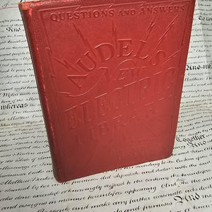 Könnte beinhalten: Ein rotes gebundenes Buch mit dem Titel Audels New Electric Library und der Prägung Questions and Answers auf dem Vorderdeckel. Das Cover zeigt ein Blitzdesign, das vom zentralen Text ausgeht, mit abgenutztem Buchrücken.
