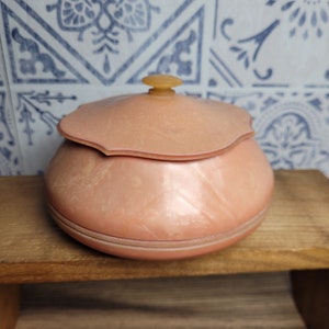 Può includere: Un contenitore vintage in plastica rosa con un coperchio. Il contenitore ha un bordo a festoni e una finitura liscia e lucida.