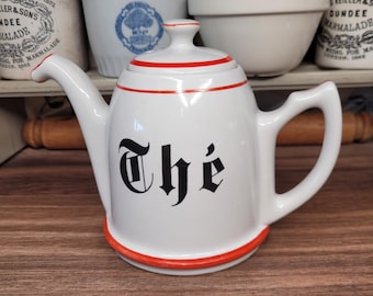 May include: White ceramic teapot with a red rim and handle, featuring the word "Thé" in black gothic lettering. The teapot has a spout and a lid with a red accent. The background includes other kitchenware.