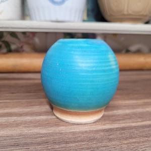 May include: A small, round, turquoise ceramic vase with a light brown base. The vase has a smooth, glossy finish and a simple, elegant design. The vase is approximately 7.6 cm in diameter and 7.6 cm tall.