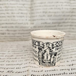 May include: A small, cylindrical white ceramic ointment pot featuring black printed text and a central illustration of a seated figure. The text reads Holloway's Ointment, with additional details and the words Trade Mark visible on the front surface.