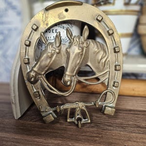 Op de afbeelding: Een messingkleurige metalen wandhaak met een hoefijzervormig frame rond de gebeeldhouwde koppen van twee paarden. Het ontwerp bevat een centrale horizontale balk met een klein haakje aan de onderkant en een verweerd metalen oppervlak.