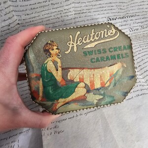 May include: Vintage Heaton's Swiss Cream Caramels tin. The tin features an illustration of a woman in a green dress, with the text "Heaton's Swiss Cream Caramels". The tin is octagonal with a decorative edge.
