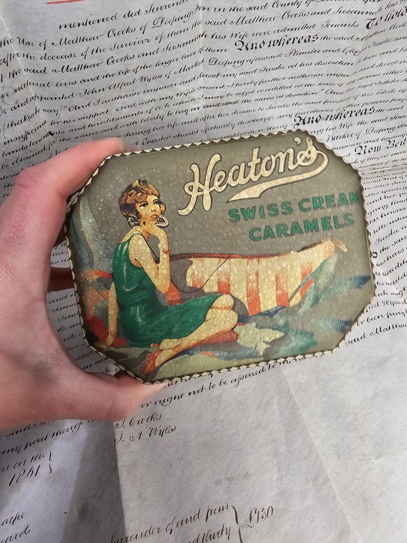 May include: Vintage Heaton's Swiss Cream Caramels tin. The tin features an illustration of a woman in a green dress, with the text "Heaton's Swiss Cream Caramels". The tin is octagonal with a decorative edge.
