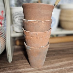 以下が含まれることがあります： 4つの小さな、風化したテラコッタ製の植木鉢のスタック。植木鉢は温かみのある赤茶色で、少しざらざらした質感です。1つの植木鉢には「BULWELL」の文字がエンボス加工されています。植木鉢は積み重ねられています。