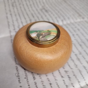 Puede incluir: Pequeña caja redonda de madera clara con tapa que presenta una pintura de paisaje en miniatura. La tapa es dorada y la pintura representa un campo verde y amarillo con árboles y un pequeño edificio.